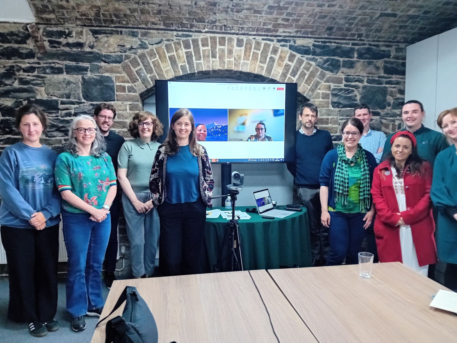 A photo of a group of people standing in front of a large screen to show 2 more people joining a meeting via Zoom. 